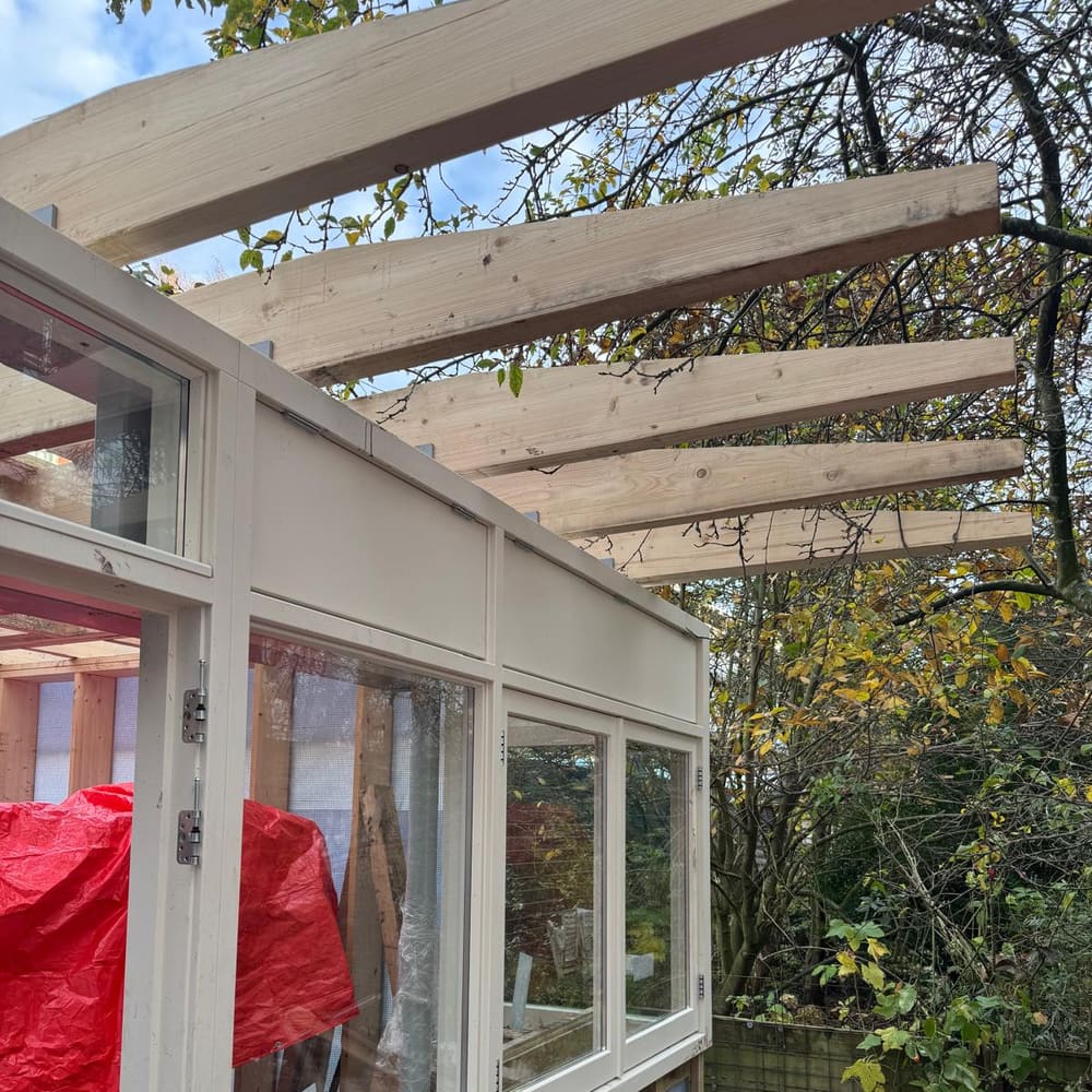 Tuinkamer in aanbouw: cr&egrave;mekleurige houten kozijnen met glas, onder een pergola van lichte balken